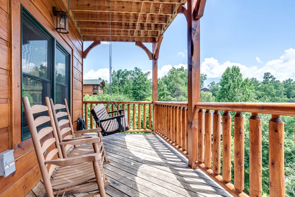 This exterior scene captures a pair of wooden rocking chairs on a porch, framed by a classic wooden railing and surrounded by lush greenery. The soft lighting suggests either sunrise or sunset, and the open-air setting connects directly to the natural landscape of the mountains.