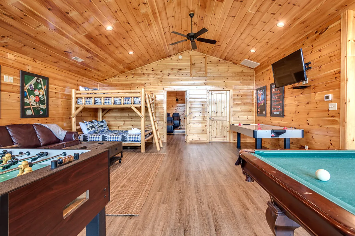 A spacious game room featuring a foosball table, pool table, bunk beds, and a leather sofa. The environment looks lively and comfortable, designed for play, relaxation, and socializing, with enough sleeping space for larger groups.