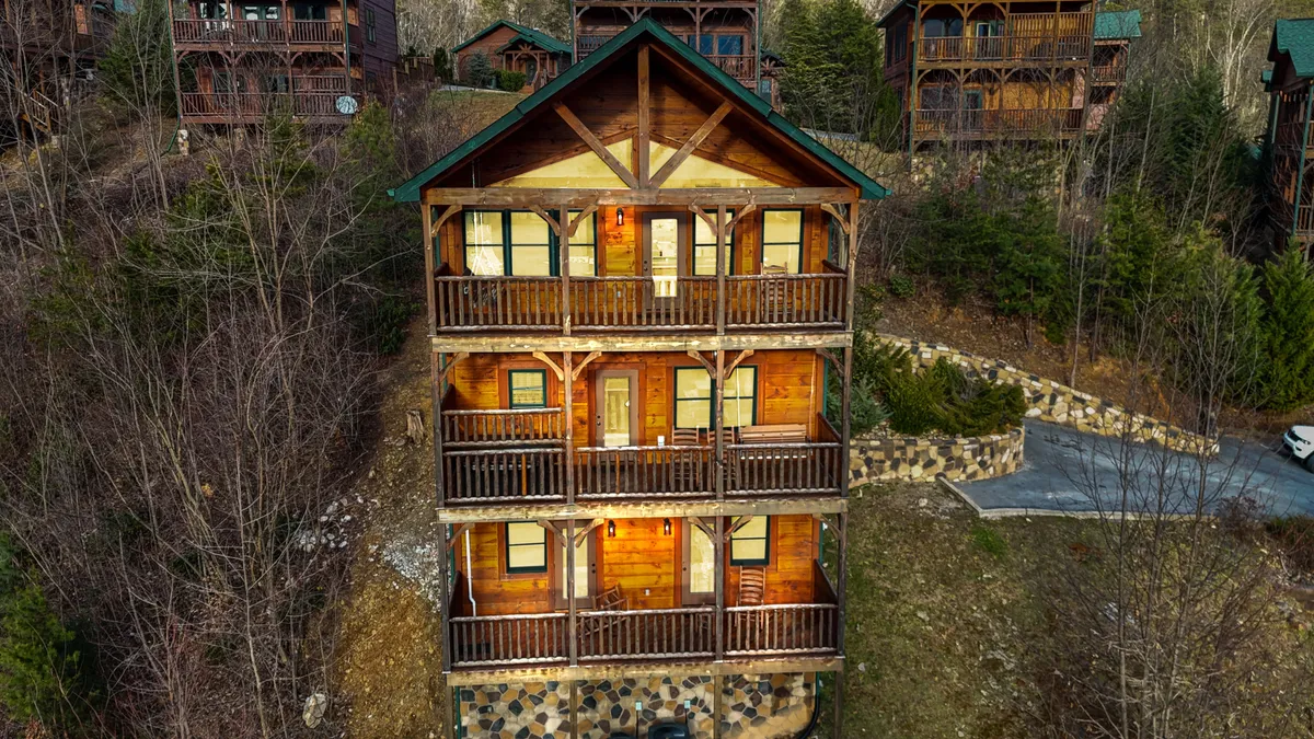 The hero image showcases the grand exterior of a three-story wooden cabin with expansive balconies overlooking the Smoky Mountains. A warm glow from inside hints at a cozy interior, while a stone pathway leads up to the inviting front entrance. The scene is bathed in sunset light, highlighting the serene and welcoming atmosphere perfect for family gatherings and outdoor relaxation.