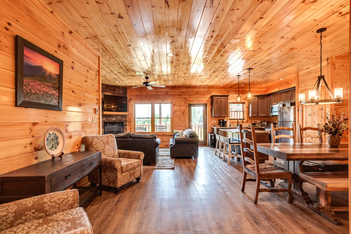 An inviting open-plan living and dining area featuring a vaulted wooden ceiling, expansive windows with mountain views, a large dining table, comfortable seating, and a fireplace. The space exudes warmth and provides a seamless flow between lounging, dining, and gathering, framed by natural light and cozy décor.