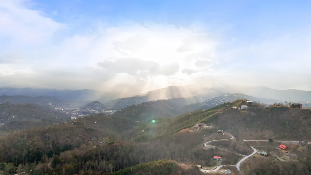 This image showcases a sweeping view of a mountain range under dramatic sunlight, with a winding road and scattered houses nestled in the landscape. The lush greenery and expansive sky evoke a sense of adventure and the natural beauty of the Great Smoky Mountains region.