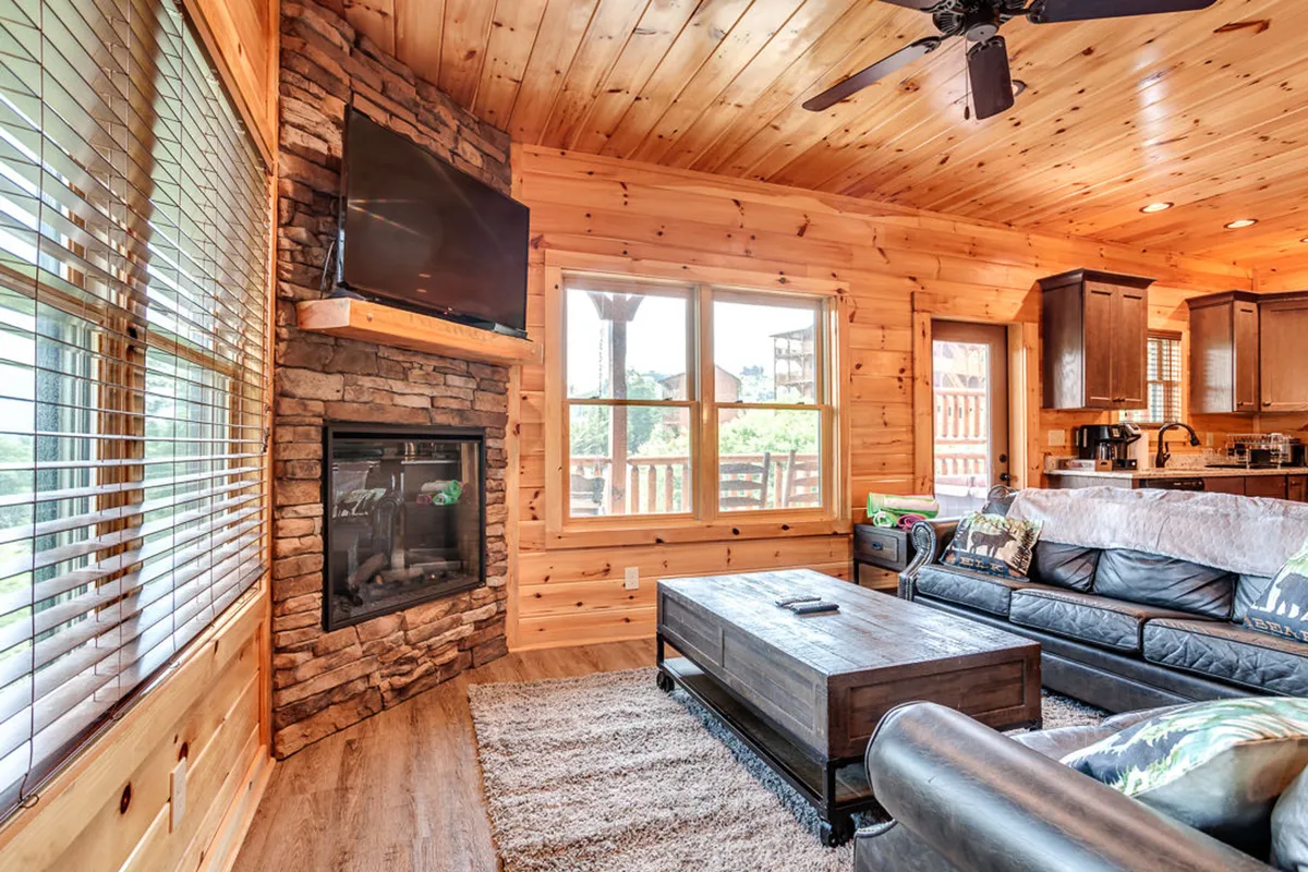 A handsome living room with a stone fireplace, large windows letting in plenty of sunlight, comfortable seating, and a coffee table. The space is ideal for relaxing, socializing, or enjoying the mountain views from inside.