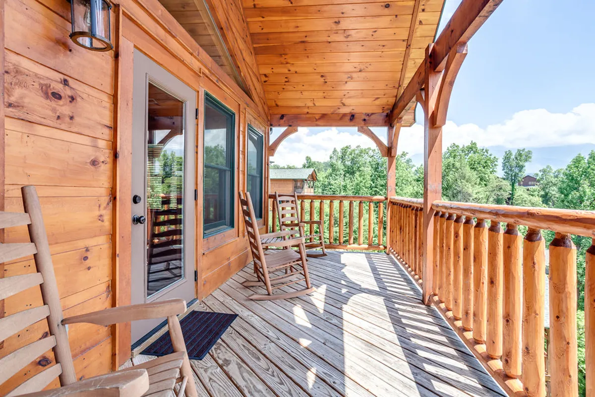 A welcoming exterior view featuring classic wooden rocking chairs on a covered porch, framed by a sturdy wooden railing and surrounded by lush natural scenery. The inviting setup suggests a peaceful spot for morning coffee, reading, or socializing while soaking in the fresh mountain air.