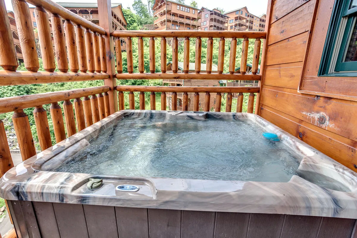 A private outdoor hot tub sits on a wooden deck, surrounded by lush greenery and protected by a sturdy wooden railing. The tranquil setting offers sweeping mountain views, perfect for relaxation in any season. The scene captures both the inviting hot tub and the serene natural backdrop.