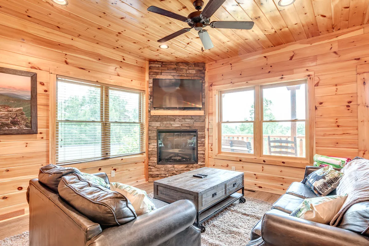 An inviting interior living area featuring a large stone fireplace, wooden ceiling and accents, leather sofas, a coffee table, and large windows that let in natural light. The space exudes warmth and is set up for group gatherings and fireside relaxation.