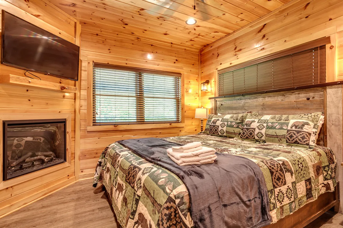 A warmly lit bedroom with wooden walls, a comfortable bed draped in patterned blankets, an electric fireplace casting a rustic glow, and a television for entertainment. The space is inviting, cozy, and perfect for relaxing after a day out.